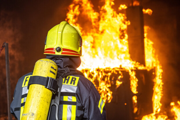 A firefighter in full gear, including a yellow helmet and air tank, stands facing a large, intense fire burning inside a structure. Bright flames and heavy smoke fill the background.