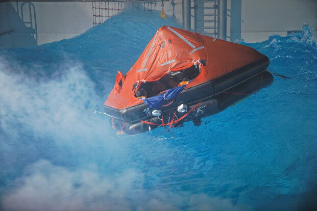 An orange inflatable life raft floats on rough blue water near a ship, with mist or spray in the foreground. The raft is partially occupied and appears to be used for an emergency or rescue scenario.