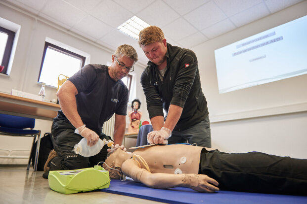 Two people practice CPR and use a defibrillator on a medical training mannequin in a classroom setting. One person gives chest compressions while the other uses a manual resuscitator mask.