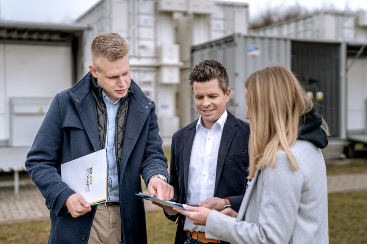 Drei professionell gekleidete Personen stehen im Freien und diskutieren über Dokumente. Ein Mann zeigt auf die Papiere, während die anderen aufmerksam zuhören. Im Hintergrund sind Industriecontainer oder -geräte zu sehen.