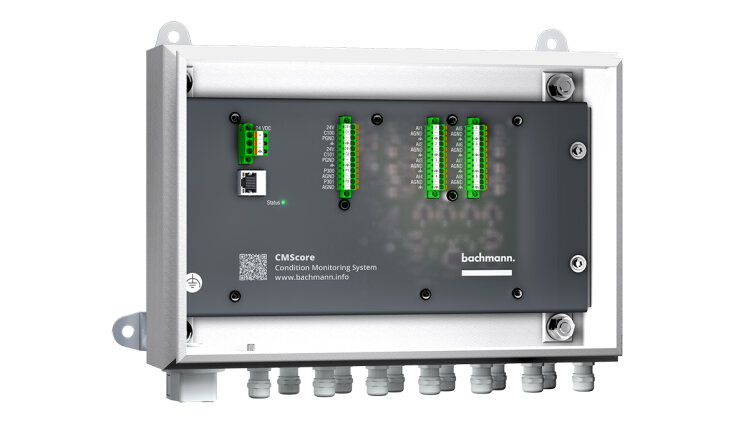A rectangular industrial monitoring device with several green and black connections, labeled CMScore Condition Monitoring System from Bachmann, is mounted on a white background. Several connections can be seen on the bottom edge.