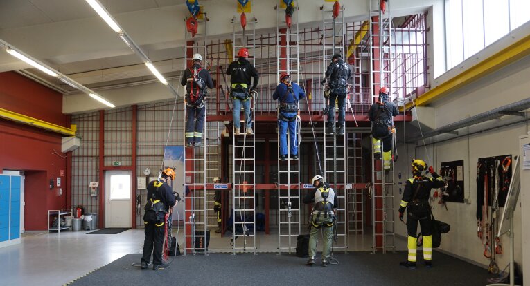 Eight people wearing helmets and safety gear practice climbing ladders attached to an indoor training structure, while two others observe; ropes and climbing equipment are visible around the area.