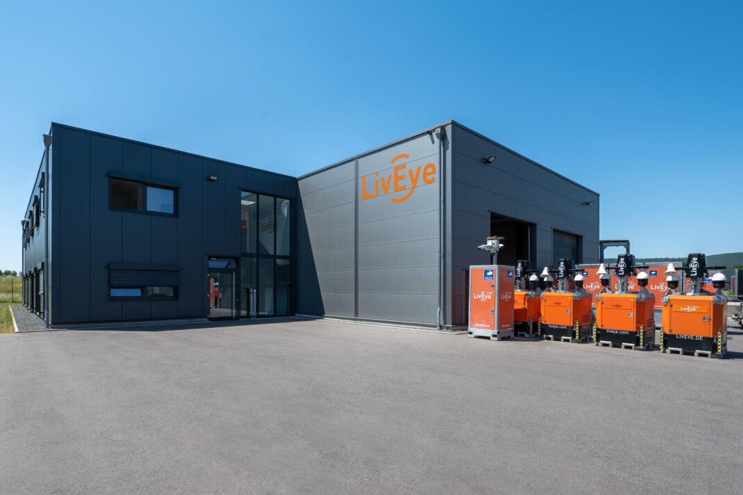 Ein modernes Industriegebäude mit "LivEye"-Schildern und mehreren orangefarbenen Sicherheitskameras auf Rädern, die an einem sonnigen Tag im Freien geparkt sind.