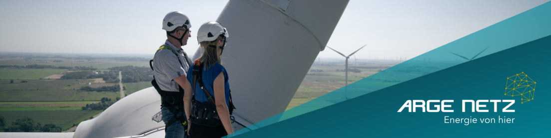 Zwei Personen in Schutzkleidung stehen auf einem Windrad und überblicken eine Landschaft mit Windrädern. Auf einem blauen Banner im Vordergrund sind das Logo der ARGE NETZ und der Text Energie von hier zu sehen.