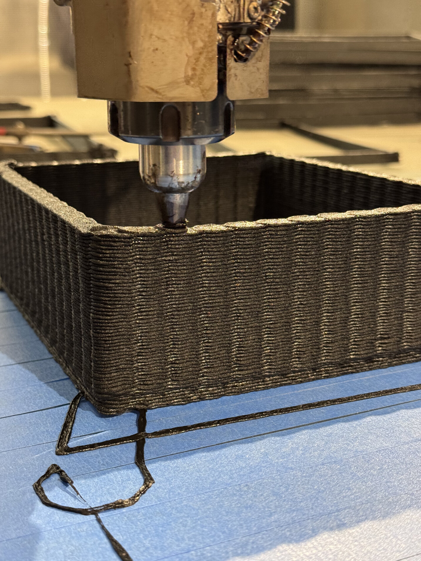 Eine Nahaufnahme einer 3D-Druckerdüse, die schwarzes Filament extrudiert, um die Wände eines rechteckigen Objekts auf einer mit blauem Klebeband versehenen Bauplatte zu formen.