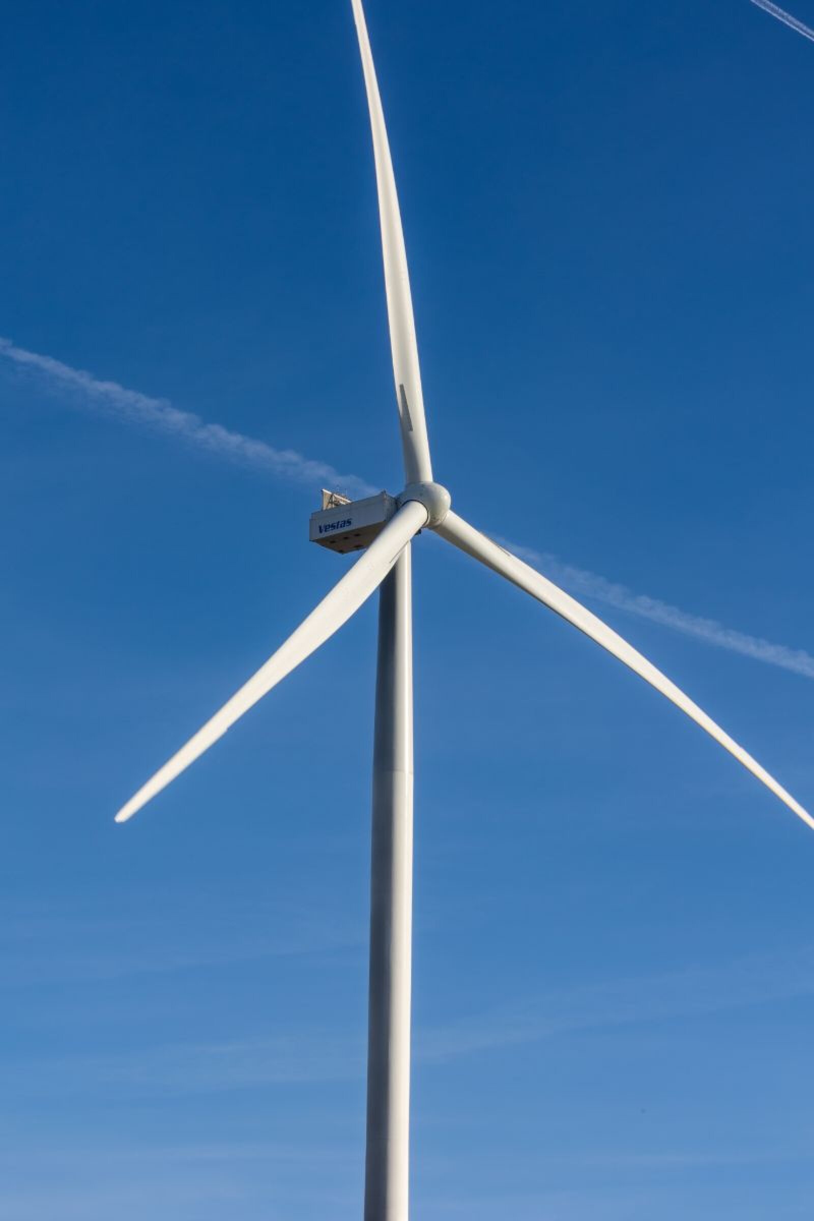 Eine hohe weiße Windturbine mit drei Flügeln steht vor einem klaren blauen Himmel, und im Hintergrund ist ein schwacher Kondensstreifen zu sehen.
