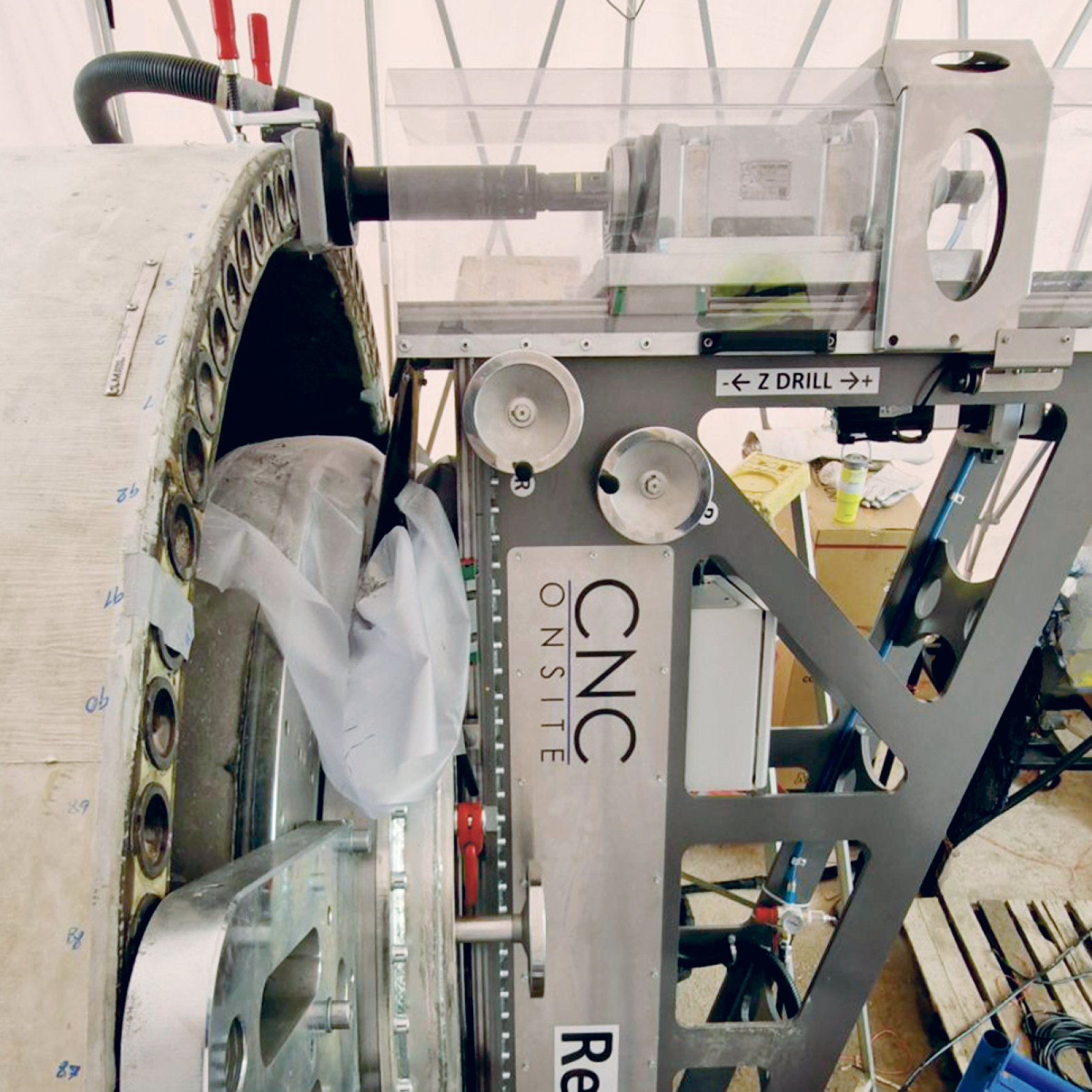Nahaufnahme einer CNC-Bohrmaschine, die an der Kante einer großen, kreisförmigen Metallstruktur, möglicherweise einer Turbinenkomponente, in einer Werkstatt arbeitet, wobei im Hintergrund Werkzeuge und Kabel zu sehen sind.