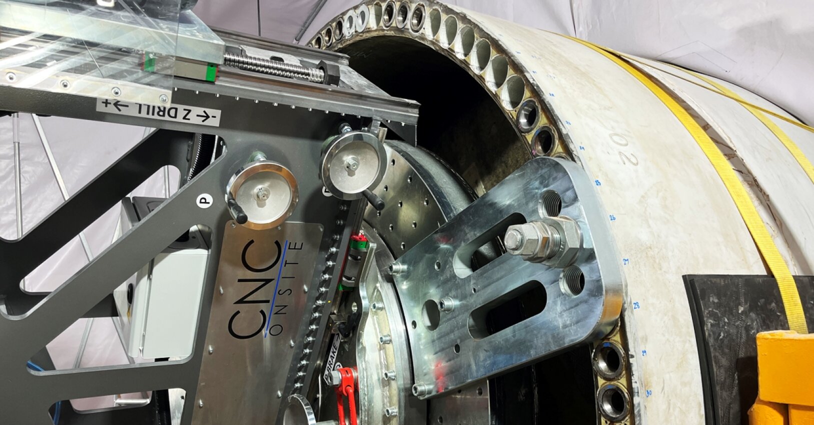 Close-up of industrial machinery from CNC Onsite working on a large cylindrical metal component, with visible gears, bolts, and various metal parts, inside a covered workspace.