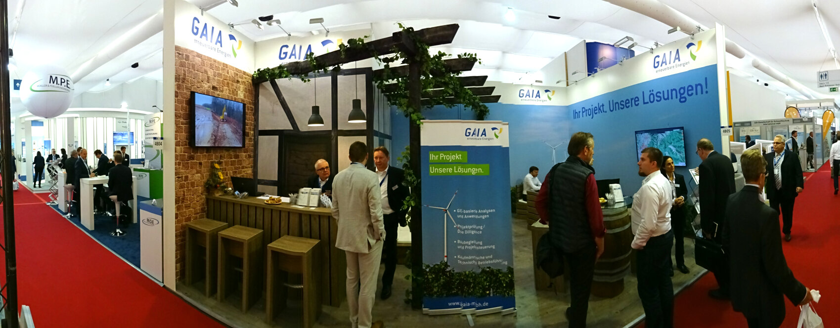 Ein Messestand mit einem GAIA-Schild, Menschen, die sich unterhalten, einem Tresen mit Hockern, Bildschirmen und Informationsbannern. Der Stand hat ein modernes Design mit Pflanzen und Holzakzenten. Im Hintergrund sind andere Stände und Besucher zu sehen.