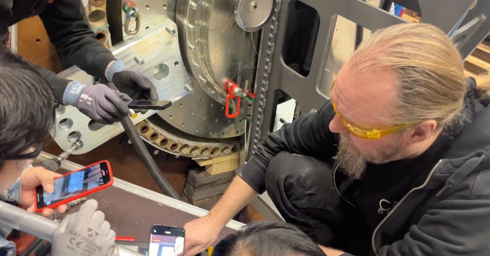 A man wearing yellow safety glasses kneels beside industrial machinery while others, wearing gloves, use smartphones to take photos and notes of the equipment.