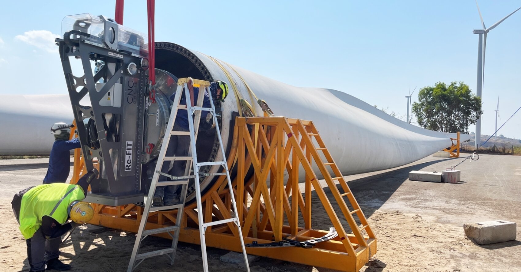Workers assemble or inspect a large wind turbine blade resting on supports outdoors, using a ladder and machinery. Two wind turbines and a tree are visible in the background under a clear sky.