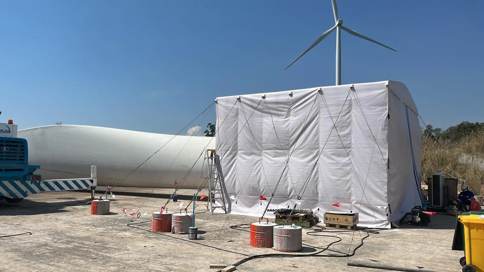 A large white tent-like structure is set up outdoors near a wind turbine blade, with equipment, cables, and weighted barrels surrounding it. A wind turbine is visible in the background under a clear blue sky.