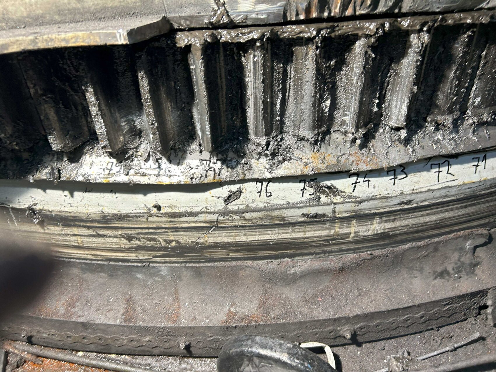 Close-up of a large, industrial metal gear coated with dark grease and grime. The gear has visible numbers marked in black ink along the edge. The surrounding area appears dirty and worn.