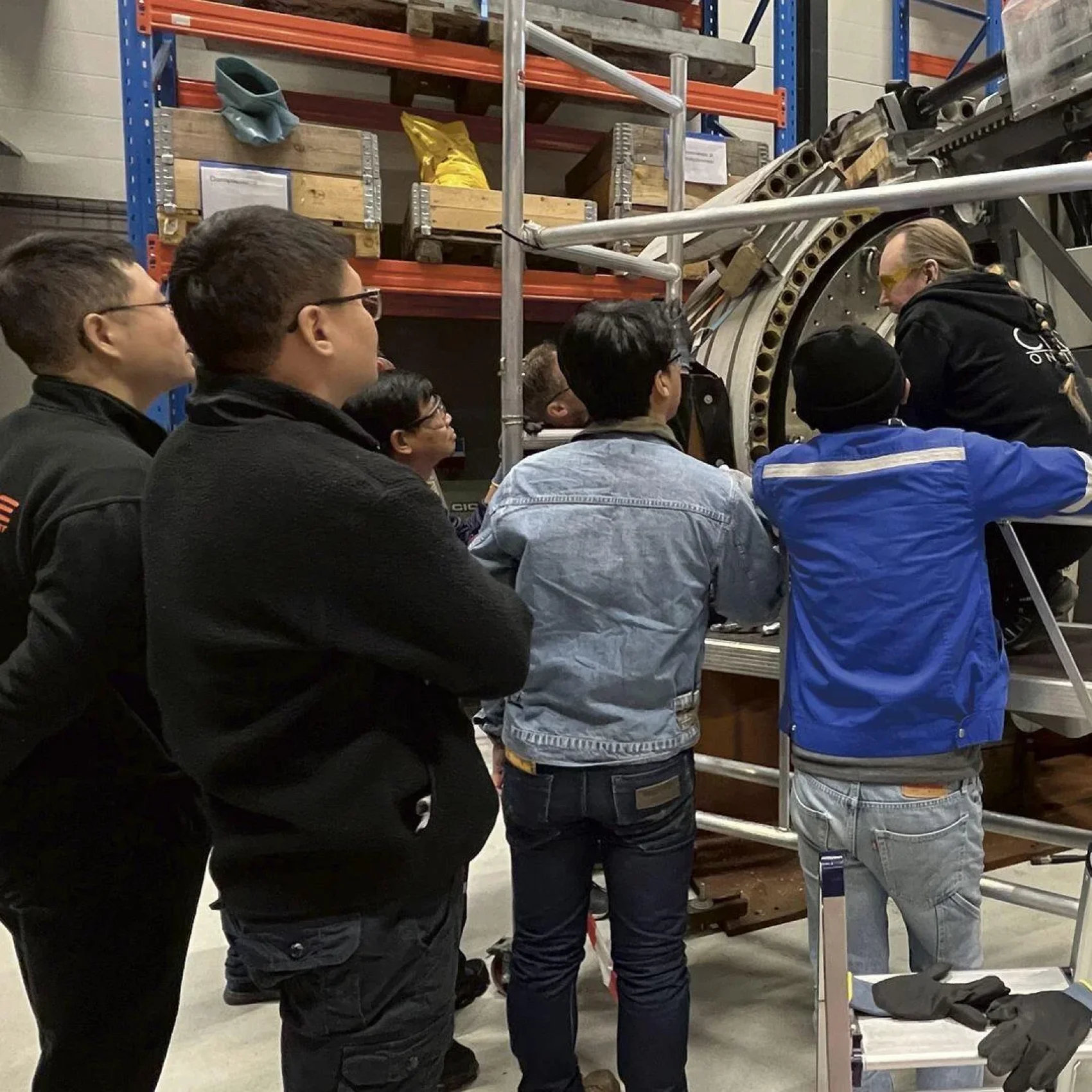 Eine Gruppe von Menschen steht in einer Industriehalle um ein großes mechanisches Bauteil herum und sieht und hört einem Ausbilder zu, der etwas in der Nähe der Maschine erklärt. Im Hintergrund sind Regale mit Kisten und Geräten zu sehen.