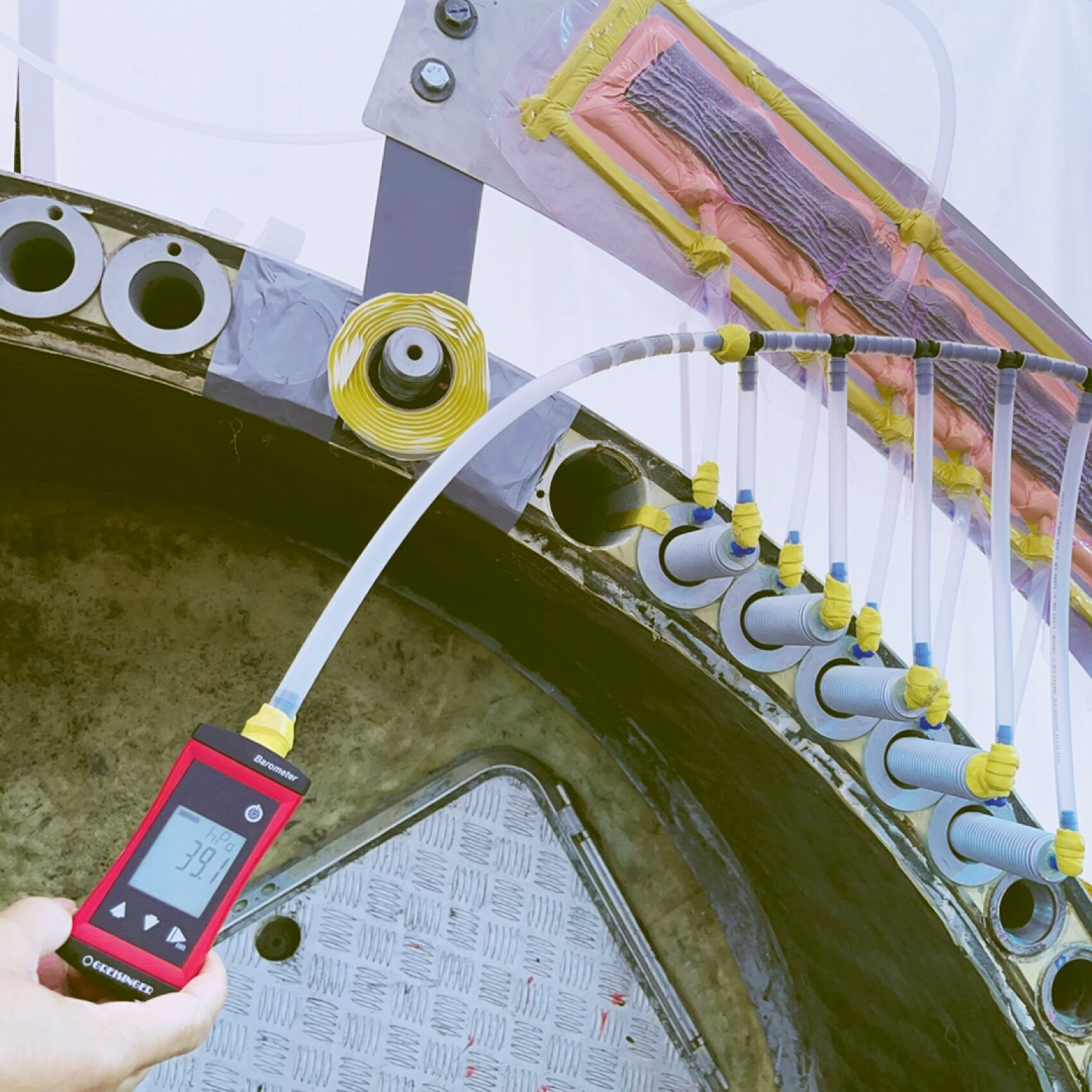 A hand holds a digital meter displaying “39.9,” connected by a tube to a large industrial apparatus with multiple pipes and cables, some wrapped in yellow tape, possibly for testing or monitoring purposes.